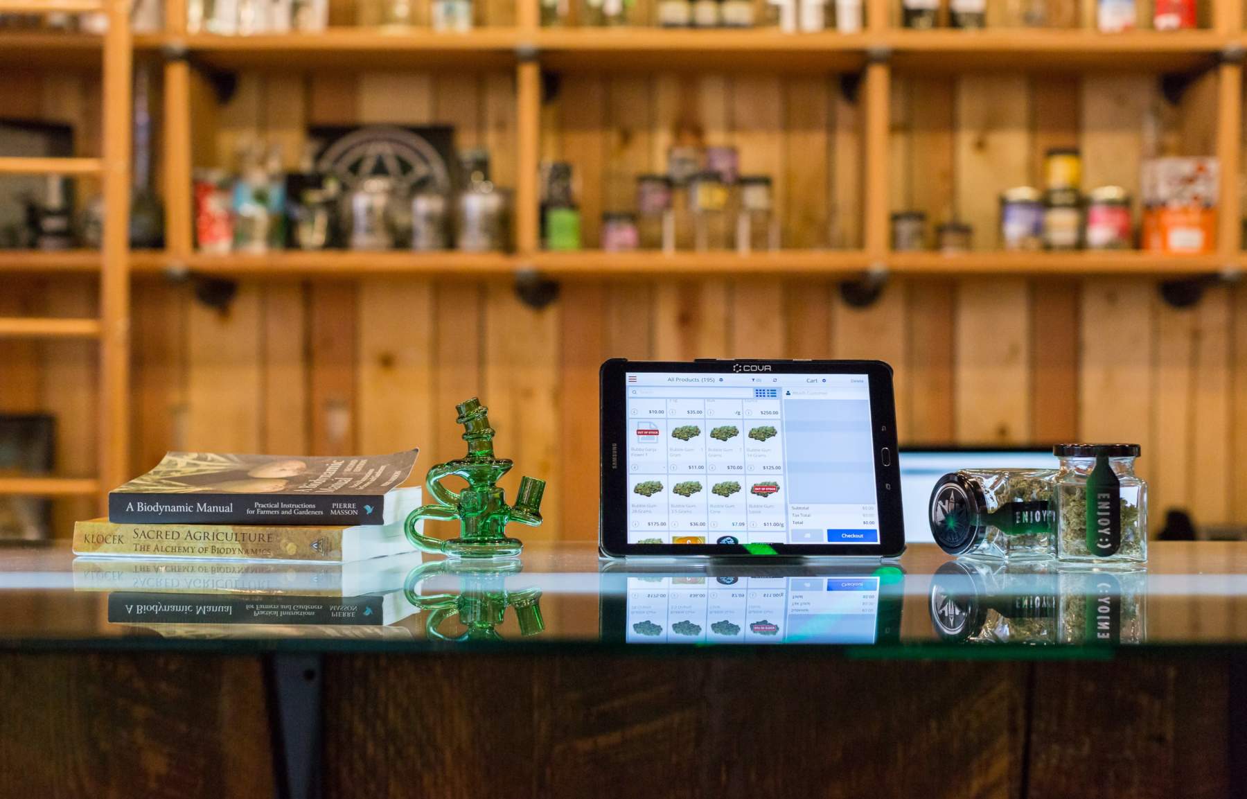 A green glass water pipe sits next to a tablet displaying cannabis products for sale, with two jars of cannabis and a stack of books on biodynamics in a rustic wooden setting.