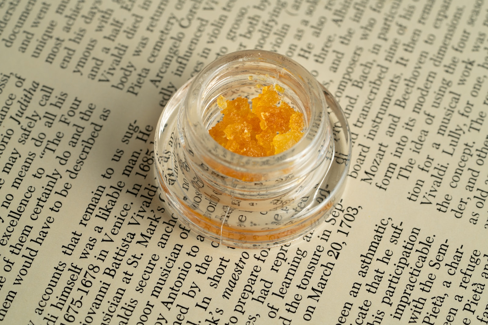 Close-up of a clear jar containing golden cannabis concentrate crystals, placed on a vintage book page filled with text. The image highlights the texture and color of the concentrate, showcasing its potential for use in various cannabis products.